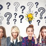 Four_young_women_sitting_near_concrete_wall_and_thinking_hard._One_of_them_has_a_good_idea._Question_marks_and_light_bulb_sketch_on_the_wall._Concept_of_brainstorming