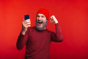 Photo_of_cheerful_man_with_white_beard_and_red_cap_looking_at_smartphone_and_celebrating_success
