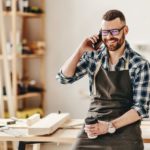 happy_young_man_in_apron_with_cup_of_takeaway_coffee_in_hand_talking_on_smartphone_while_leaning_on_workbench_in_modern_workshop_