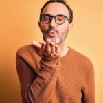 Middle_age_hoary_man_wearing_brown_sweater_and_glasses_over_isolated_yellow_background_looking_at_the_camera_blowing_a_kiss_with_hand_on_air_being_lovely_and_sexy._Love_expression.