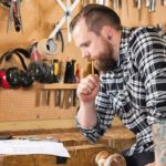 Adult_craftsman_sits_and_looks_at_plans_in_a_work_shop_for_wood_work_and_construction._Carpenter_designs_and_prepares_new_work._