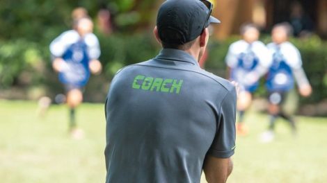 Male_soccer_or_football_coach_in_gray_shirt_with_word_COACH_written_on_back,_standing_on_the_sideline_watching_his_team_play,_good_for_sport_or_coaching_concept