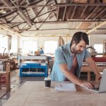 Young_woodworker_with_a_beard_leaning_over_a_workbench_in_his_large_workshop_full_of_carpentry_equipment_working_online_with_a_laptop