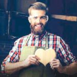 Happy_handsome_joiner_in_love_holding_and_showing_wooden_heart_near_tabletop_with_tools.__Stylish_young_entrepreneur_with_beautyful_hairstyle_and_saved_glasses_smile_at_his_workstation._He_love_his_job